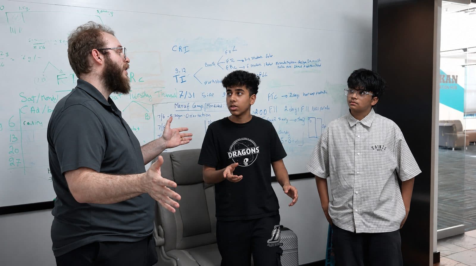A mentor at a whiteboard covered in planning notes, working through a problem with two ITKAN students