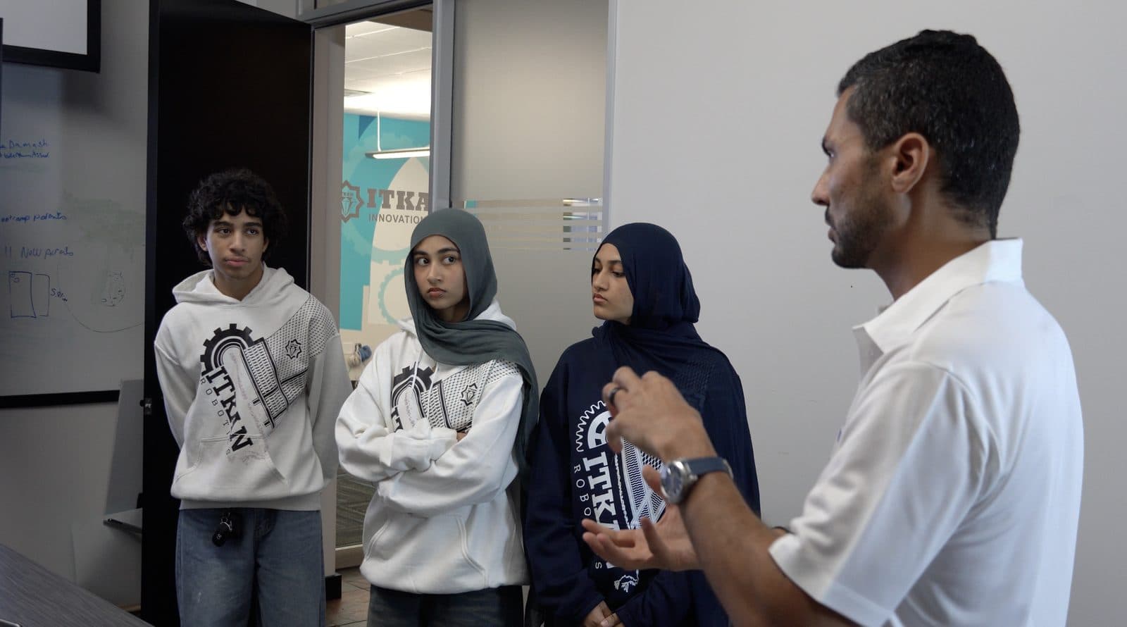Three ITKAN students in team hoodies listening as Ashraf Ahmed talks them through a point