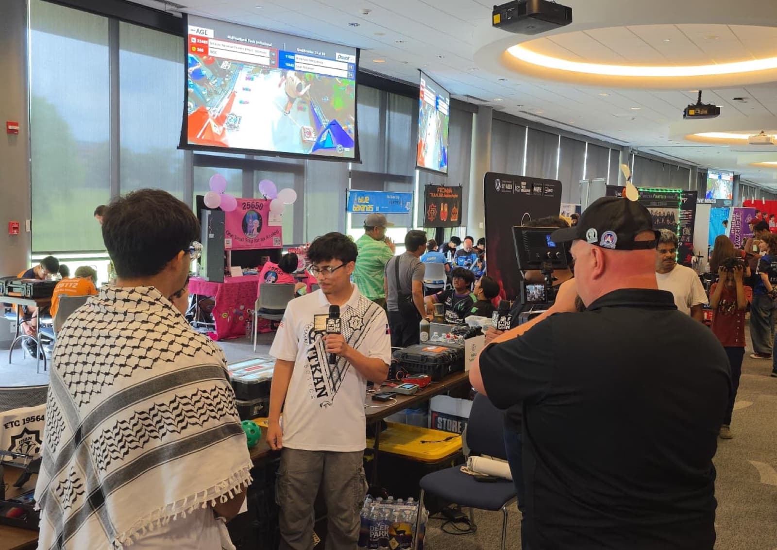 An ITKAN Solar student holds a microphone for an on-camera interview as a videographer films, in the venue at the Multinational Tech Invitational