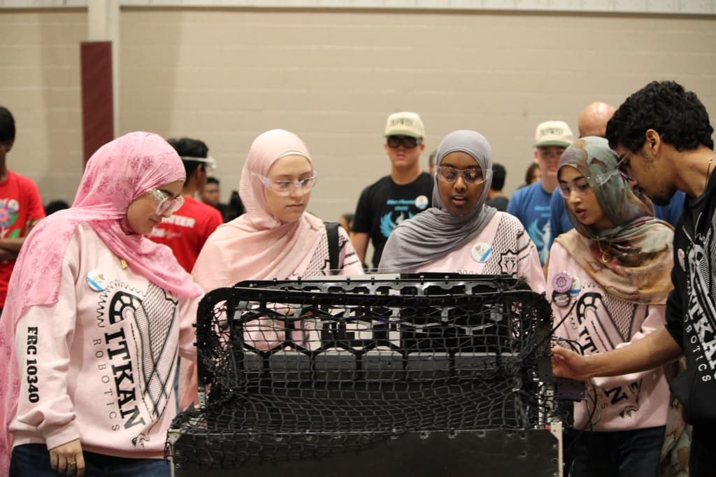 Four 10340 team members in safety glasses and pink ITKAN hoodies gather around the robot with Coach Adam